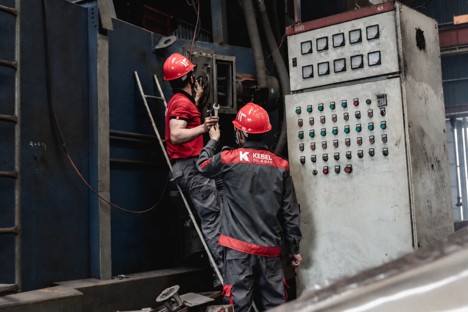 Dos técnicos trabajan en maquinaria industrial; fotografía ilustrativa con el logotipo de Kebel añadido digitalmente al uniforme